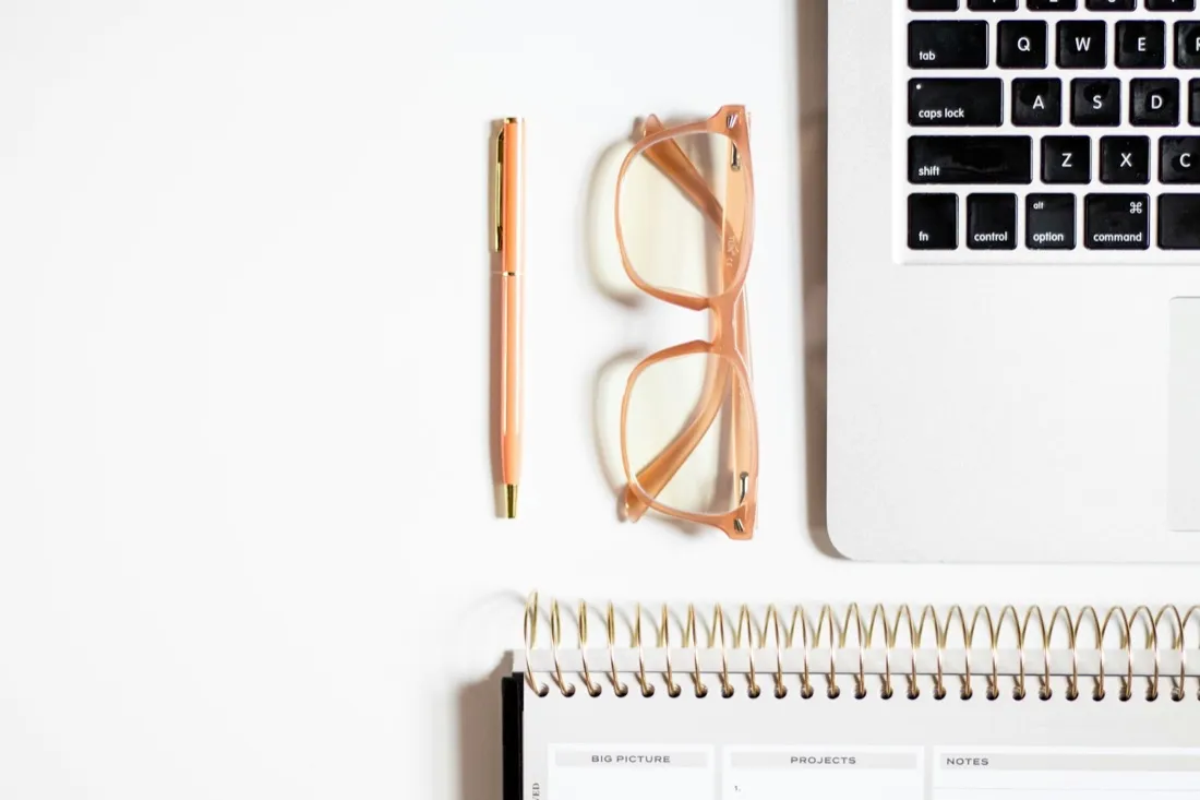 A laptop, glasses and a notepad arranged on a desk
